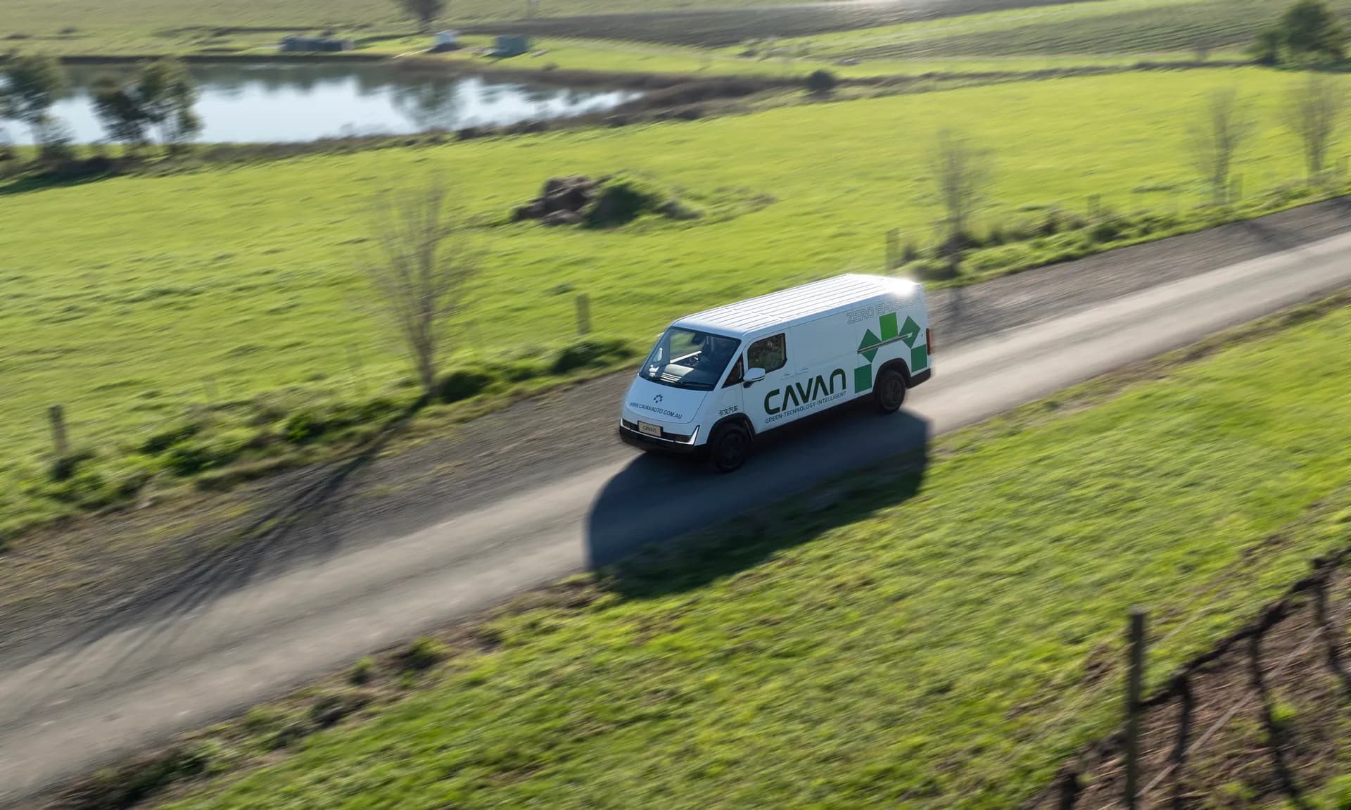Aerial view of a Cavan electric van driving a country lane past a farm dam